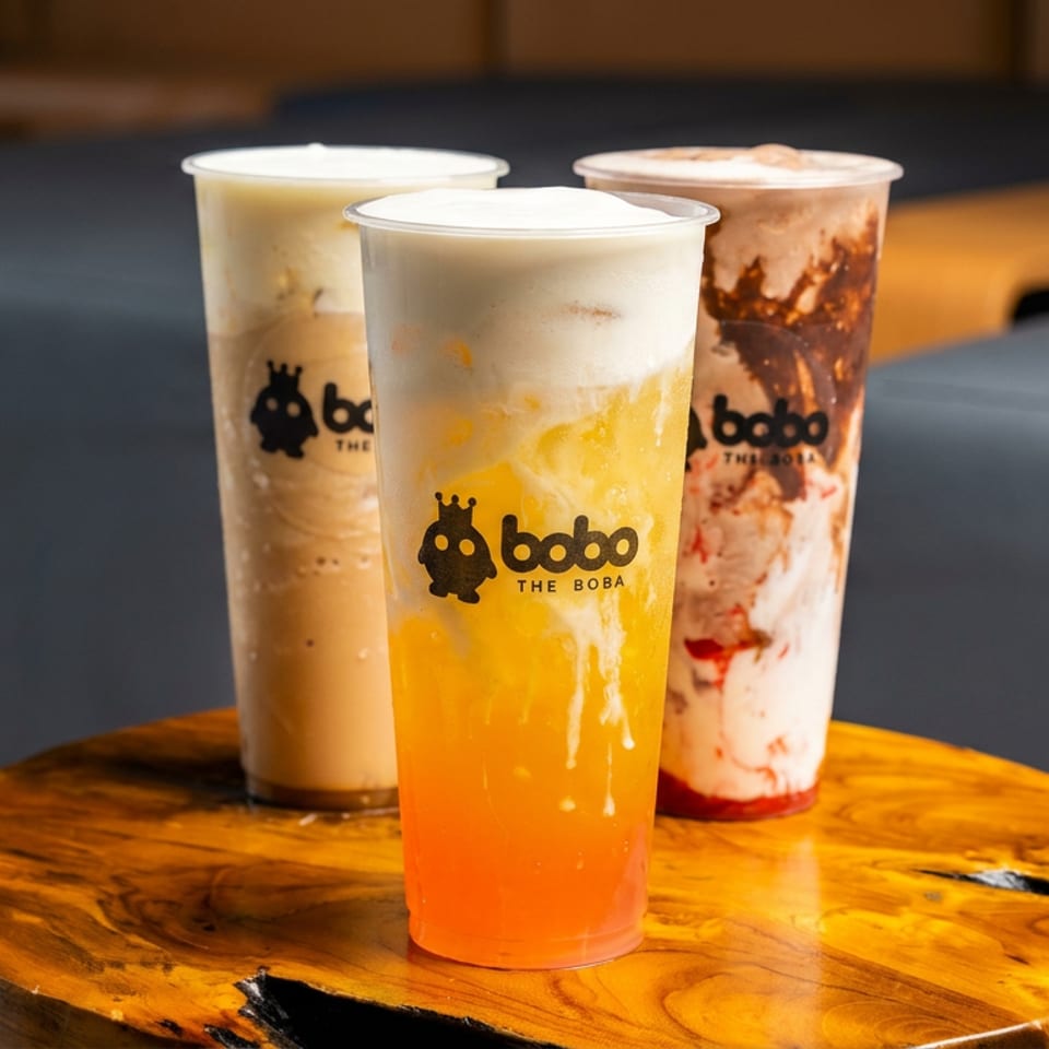 Three Bobo The Boba drinks on a wooden table: a brown drink, a yellow drink with foam, and a red and white swirled drink.